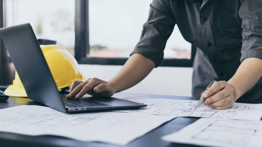 Automation engineer sedang menganalisa blueprint arsitektur sistem kontrol dan bekerja menggunakan laptop